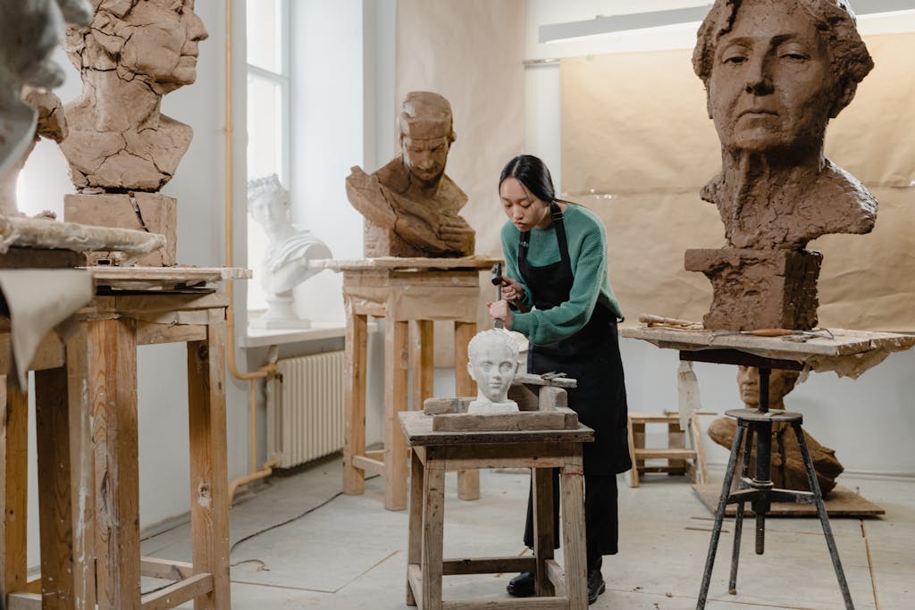 A female sculptor meticulously works on a clay bust in a well-lit art studio filled with large sculptures.